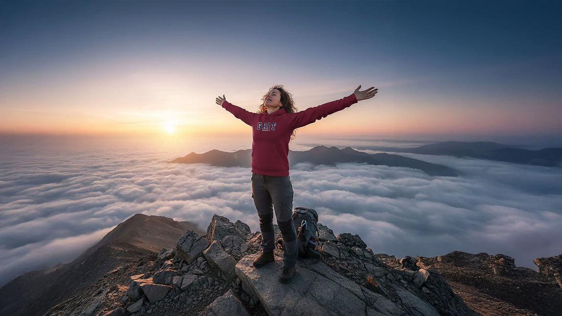 Photo of a person wearing a maroon 'GRIT' hoodie standing on a mountain peak above clouds at sunrise, celebrating personal achievement, freedom and success, symbolizing perseverance and personal triumph, reflecting the core values of the Wear Grit Brand