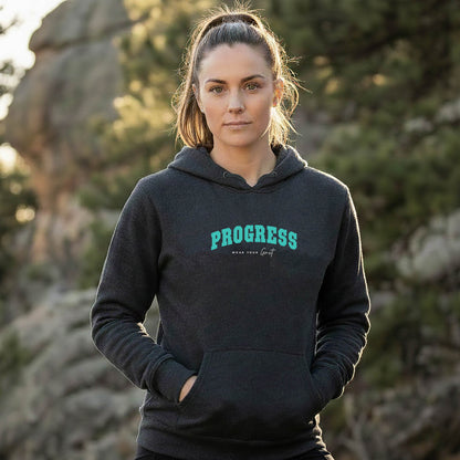 A female athlete wearing a black hoodie with PROGRESS print, standing outdoors with trees in the background. From wear grit brand clothing