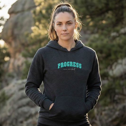 A female athlete wearing a black hoodie with PROGRESS print, standing outdoors with trees in the background. From wear grit brand clothing