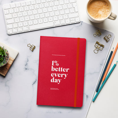 Red notebook with '1% better every day' on a marble desk with office supplies. Growth Mindset & Productivity Journal For Speaker, leaders, executives, professionals and business owners by wear grit brand