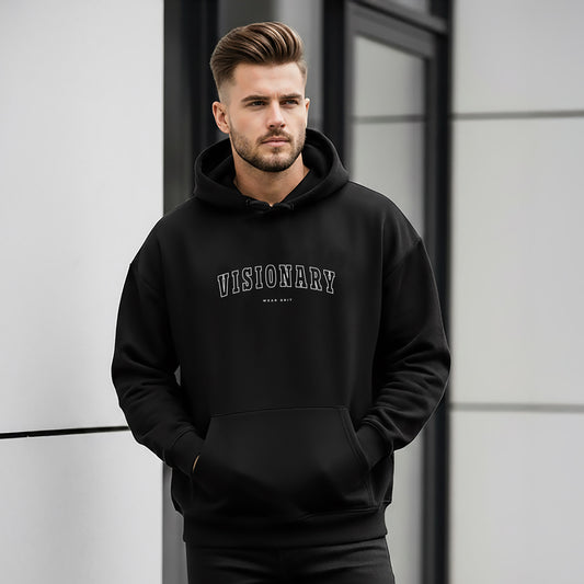 A man wearing a black hoodie with 'VISIONARY' text, standing against a modern building, Fashion hoodie with affirmation print – Visionary Hoodie, Inspirational apparel, comfort hoodie for growth mindset